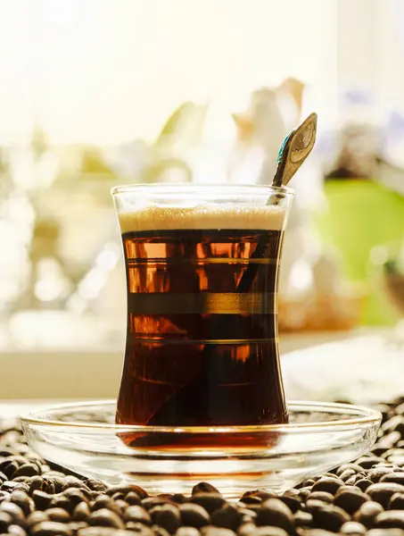A glass mug in the traditional Turkish style with black coffee stands on coffee beans lying on the table. A delicious energy drink on the background of the kitchen