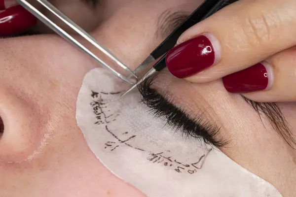 Eyelash extension procedure. Master tweezers sets fake lashes on beautiful woman close-up.