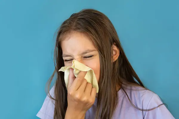 cold girl blows her nose in a white rag on blue background. Influenza or Rhinitis snot runny nose stuffy nose. Allergy young girl holding handkerchief. Medical concept.