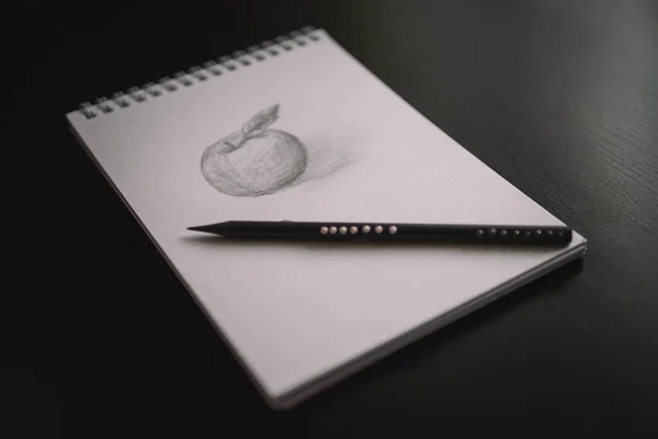An apple pencil drawing in a notebook and a pencil on a black table. Learning to draw a concept.