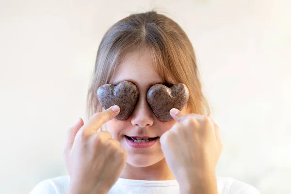 Close up glad joyful laughing child closing eyes by two cookies pastry in shape of heart in her hands, indulges. Little girl posing on white background. Concept Easter holiday baking