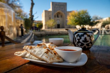 samsa with tomato sauce in a white plate, fruit teapot of tea on the background of a madrasah. Central Asian cuisine clipart