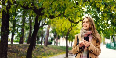 Elinde akıllı telefonuyla parkta yürüyen genç ve güzel bir kadın. Telefonda konuşmak ya da dışarıda mesaj göndermek.