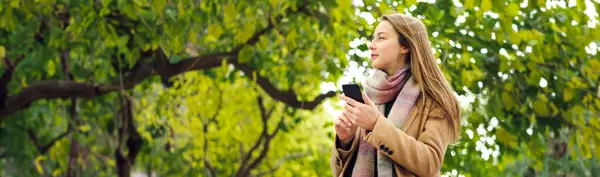 Elinde akıllı telefonuyla parkta yürüyen genç ve güzel bir kadın. Telefonda konuşmak ya da dışarıda mesaj göndermek.