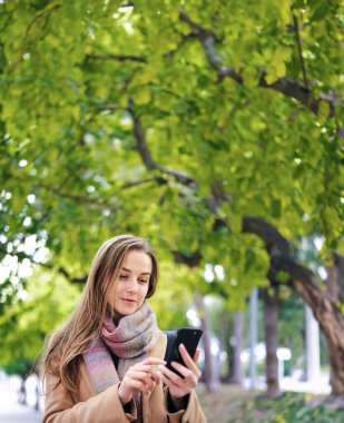 Elinde akıllı telefonuyla parkta yürüyen genç ve güzel bir kadın. Telefonda konuşmak ya da dışarıda mesaj göndermek.