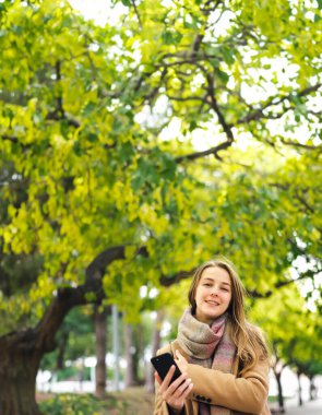 Elinde akıllı telefonuyla parkta yürüyen genç ve güzel bir kadın. Telefonda konuşmak ya da dışarıda mesaj göndermek.