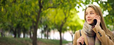 Elinde akıllı telefonuyla parkta yürüyen genç ve güzel bir kadın. Telefonda konuşmak ya da dışarıda mesaj göndermek.