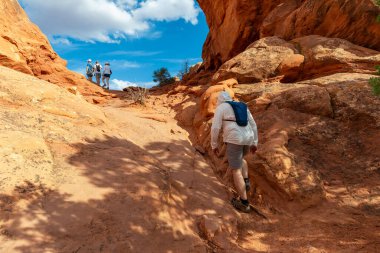 ABD 'nin Utah eyaletindeki Arches Ulusal Parkı' ndaki Windows Section Trail 'de dağ yürüyüşüne çıkan turistler. Beyaz Güneş Koruyucu Giysi ve Su Sırt Çantası Giyen Turist