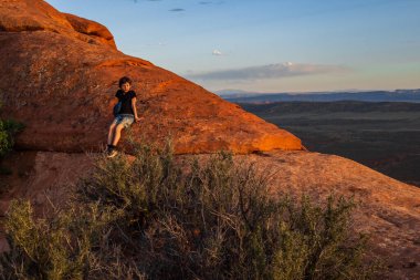 Utah Arches Ulusal Parkı 'nda gün batımında Şeytanların Bahçesi Yolu' nda yürüyüş yapan genç bir çocuk. Altın saat ışığı, kızıl kaya manzarasını yıkıyor. Geniş çöl manzaraları için çarpıcı bir zemin oluşturuyor..