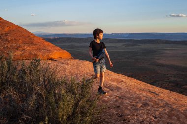 Utah Arches Ulusal Parkı 'nda gün batımında Şeytanların Bahçesi Yolu' nda yürüyüş yapan genç bir çocuk. Altın saat ışığı, kızıl kaya manzarasını yıkıyor. Geniş çöl manzaraları için çarpıcı bir zemin oluşturuyor..