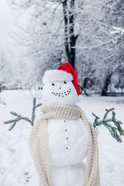 Happy snowman made outdoors in snowy winter park dressed in Santa hat, scarf with fir branches in hands. Christmas fun activities