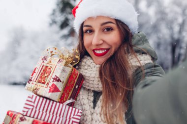 Karlı kış parkında elinde Noel hediyeleriyle selfie çeken bir kadının portresi. Bayram şenliği. Noel Baba şapkalı ve kırmızı rujlu bir kız.