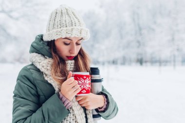Karlı kış parkında elinde elektrikli süpürge şişesiyle çay içen genç bir kadın. Kırmızı örgü Noel süveteri giymiş bir kupa. Açık havada yürüyorum. Boşluk