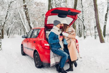 Sevgili çift arabanın bagajında oturmuş, karlı kış ormanlarında sıcak çay içiyorlar. Tatillerde insanlar dışarıda dinleniyor.