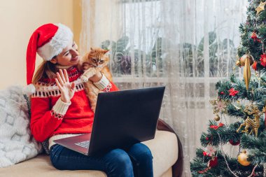 Kediyle Noel kutlaması. Noel Baba 'nın şapkalı kadın evdeki yılbaşı ağacının yanında dizüstü bilgisayar kullanıyor. İnternetteki görüntülü sohbetteki aileyi arayıp selam vermek