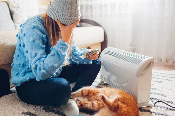 stock image Expensive electricity. High prices for heating. Using heater at home in winter with cat. Sad woman holding money wearing warm clothes