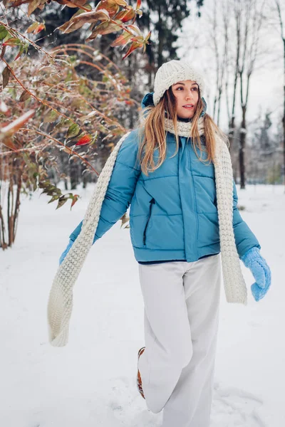 Full body portrait of young woman walking in snowy winter park wearing blue coat, knitted warm clothes. Female fashion for cold weather