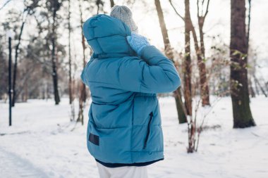 Karlı kış parkında, yansıtıcı şeritli mavi ceketli, bantlı bir kadının arka görüntüsü. Soğuk hava için sıcak kıyafetler.
