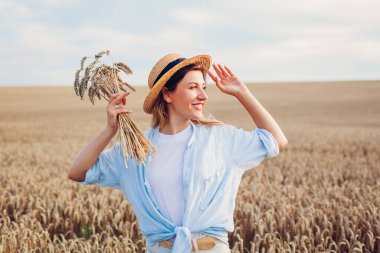 Portrait of smiling young woman walking among wheat in summer field wearing straw hat holding bundle of ripe wheat at sunset. Free girl enjoys life