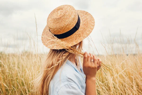 Back view of young woman walking among high grasses in summer meadow wearing straw hat and linen shirt picking bundle of dry grass. Harmony and balance