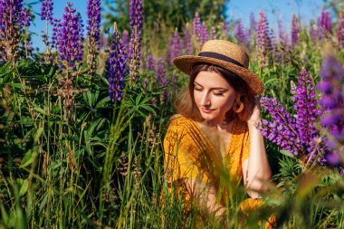 Çiçekli yaz çayırlarında dinlenen genç bir kadın elinde lupin çiçekleri buketi tutuyor. Hasır şapka giyen şık bir kız. Moda kadın aksesuarları.