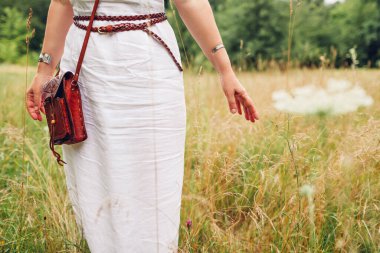 Boho style fashion. Woman wearing white linen dress, jewelry, carrying leather handmade handbag outdoors walking in summer meadow among grasses