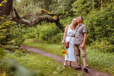 Stylish senior family couple walking outdoors in summer forest. Elderly people holding hands. Woman carries bouquet of flowers. Man and woman enjoy landscape