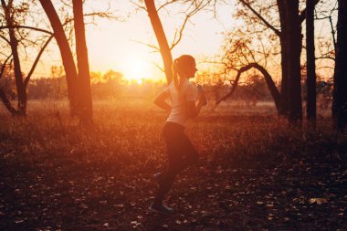 Sonbahar parkında genç koşucu eğitimi. Gün batımında su şişesiyle çalışan bir kadın. Aktif yaşam tarzı. Zayıf, sağlıklı bir kızın silueti.