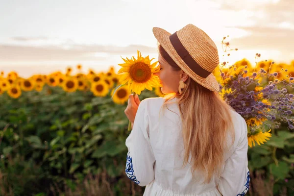 Back view of woman enjoying view in blooming sunflower field at sunset with bouquet of flowers. Girl wearing white dress and straw hat. Space