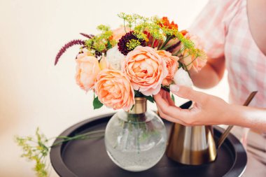 Close up of fresh english roses Lady of Shallott. Woman makes bouquet in vase of orange red flowers with greenery on table at home.