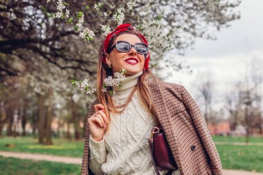 Bahar parkında çiçek açan kızıl saçlı atkı ceket ve güneş gözlüğü takan kadın portresi. Retro kadın modası. Parlak başörtüsü ve çanta. Saç stilleri için şık aksesuarlar