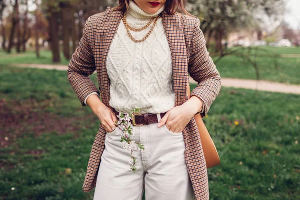 Outdoor close up of young woman wearing white sweater and pants with brown blazer holding purse. Retro style jewellery accessories and clothing. Female spring fashion