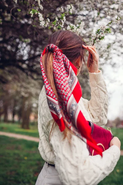 Back view of stylish young woman wearing red hair scarf in spring park by blooming trees holding purse. Retro female fashion. Bright accent headscarf for ponytail. Hair accessories