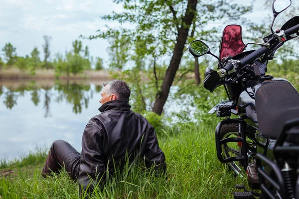 バイクで屋外でリラックスするシニアバイカーのバックビュー 川の風景を楽しむ革のジャケットを着て男 夏を保証する高齢者のオートバイ旅行 — ストック写真
