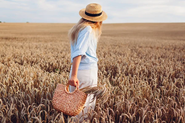 Back view of young stylish woman in hat walking in summer field holding straw handbag with wheat bundle at sunset. Harmony and freedom