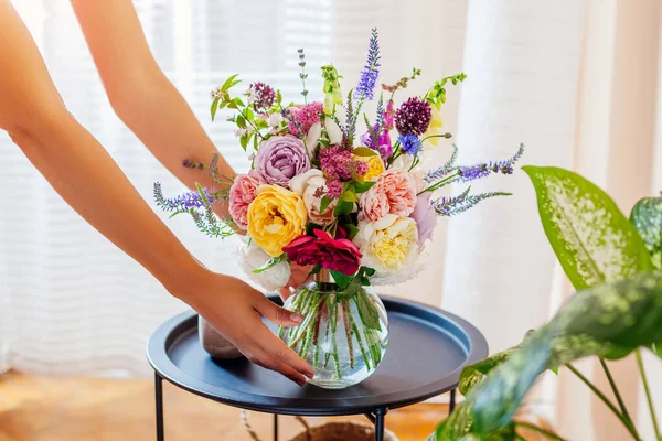 Woman puts transparent vase with bouquet of roses flowers on table. Taking care of interior and summer decor at home. Close up