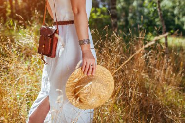 Kırsal boho tarzı. Beyaz elbise, mücevher, deri çanta ve çayırda yürüyen hasır şapka giyen şık bir kadın.