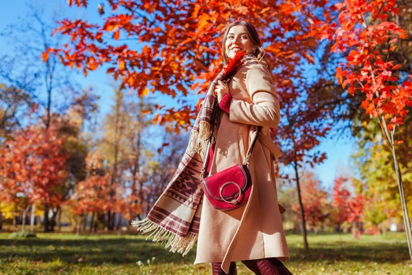 Happy young woman walking in autumn park among red trees. Female fall fashion. Stylish girl wearing beige coat and holding purse