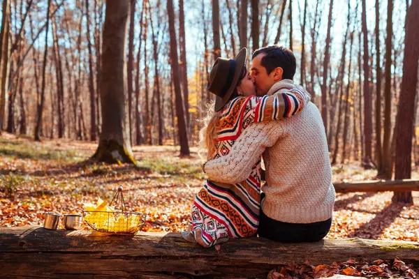 Back view of couple hugging in autumn forest. Man and woman sitting on tree trunk enjoying fall landscape on date. Young people kissing