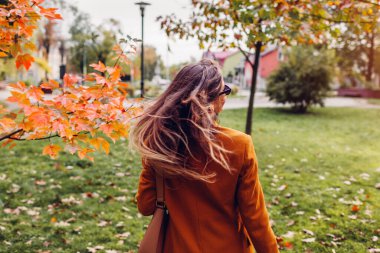 Sonbahar parkında elinde çantasıyla turuncu ceket giyen şık bir kadının arka görüntüsü. Dalgalı saçlı genç bayan sonbahar ağaçlarının arasında yürüyor..