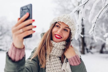 Karlı kış parkında akıllı telefondan selfie çeken genç bir kadının portresi. Sıcak kıyafetler giyen ve kırmızı rujlu bir kız manzaranın tadını çıkarıyor. Kapat.