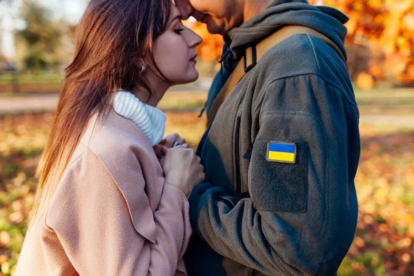 Close up of couple holding hands. Sad woman hugging husband in ukrainian military uniform with flag chevron. Upset patriotic couple outdoors. Back from war