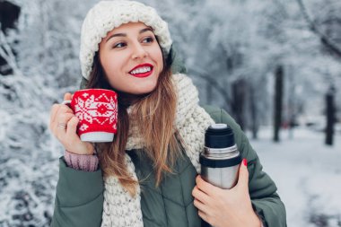 Karlı kış parkında elinde elektrikli süpürge şişesiyle çay içen bir kadın, karlı manzaranın tadını çıkarıyor. Kırmızı örgü Noel çantası giymiş bir fincan.