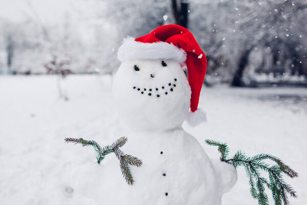 Close up of happy snowman wearing Santa hat in snowy winter park with fir branches at Christmas season outdoors. Winter activities