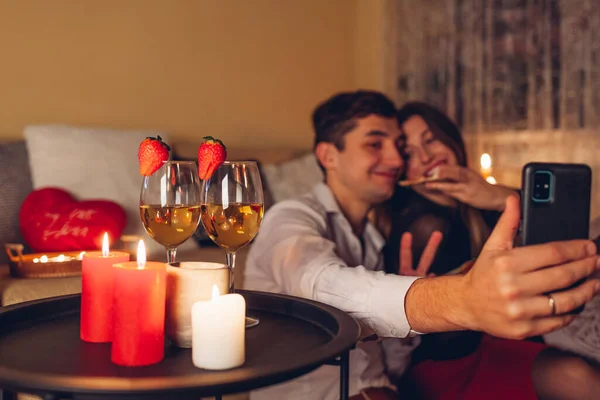 Valentines Day loving couple taking selfie on phone by wine glasses with candles having pizza romantic dinner at home. Happy man and woman celebrating at home at night