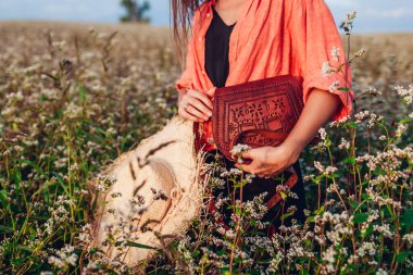 Kahverengi deri çanta ve kadın ellerindeki hasır şapkayı kapat. Gün batımında çiçeklerin arasında çiçek tarlasında aksesuar tutan bir kadın. Boho tarzı çanta.