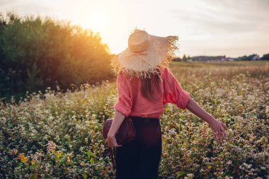 Hasır şapkalı şık bir kadın günbatımında elinde deri çantasıyla çiçek açmış karabuğday tarlasında yürüyor. Doğayla uyum içinde.
