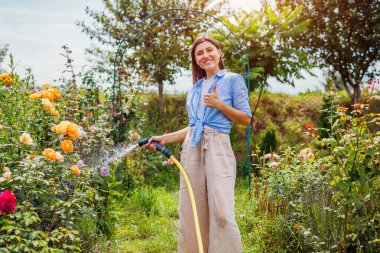 Cheerful woman gardener watering plants with hose pipe in summer garden showing thumb up. Taking care of blooming flowers enjoying landscape. Outdoor activity clipart