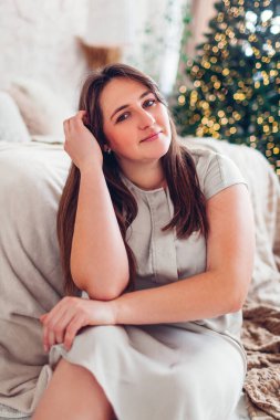 Genç bir kadının portresi. Evde Noel ağacının yanında, yatak odasında elbise giymiş poz veriyor. Dekorasyonlu, rahat bir tatil merkezi.