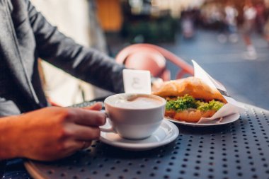 Bir fincan kahveyi erkek ellerinde kruvasanla kapat. Gün batımında açık kafede sıcak kapuçino içen adam terasta masada oturuyor..